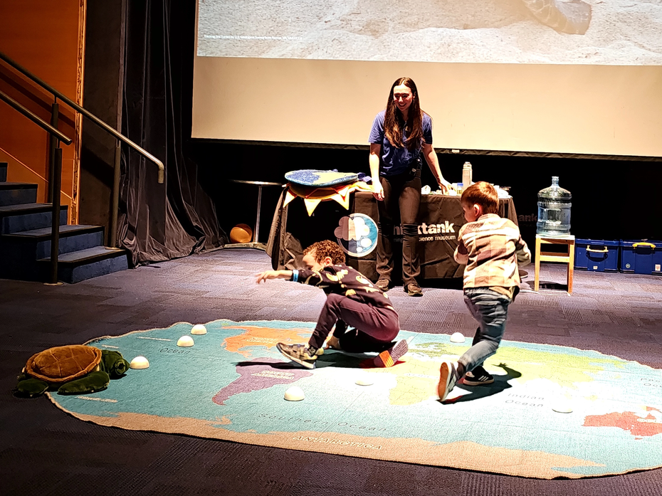 Science communicator running a session with two children on a floor mat with toy tortoise